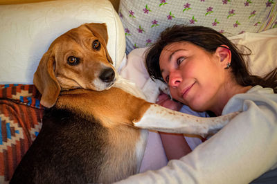 A woman lying on the bed observing her beagle puppy dog looking curiously towards the camera