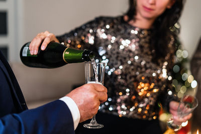 Unrecognizable female in elegant shiny dress standing in room and pouring cold champagne in glass of man during christmas party