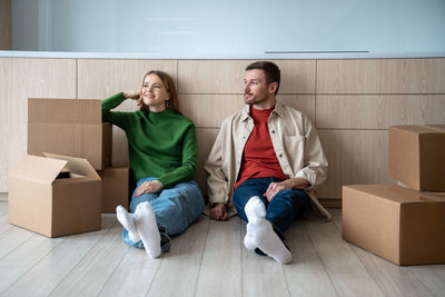 Tired glad couple sitting on floor in own new apartment, resting after carrying cardboard boxes
