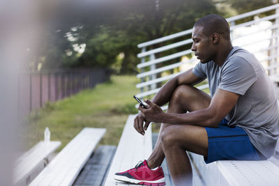 Side view of man using mobile phone while sitting on bleacher at park