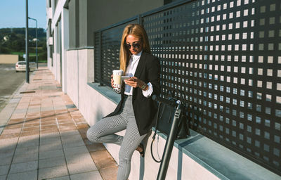 Businesswoman using mobile and drinking coffee sitting on a wall with her scooter next