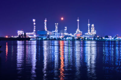 Illuminated factory by sea against sky at night oil refinery