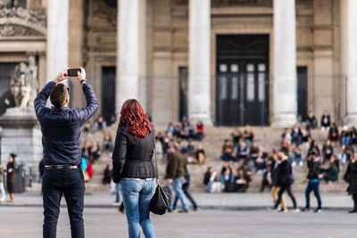 Rear view of man photographing by friend standing against historic building