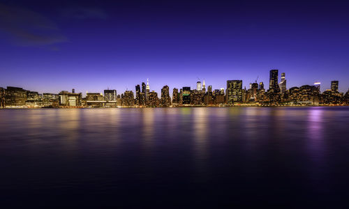 Illuminated buildings by river against blue sky at night