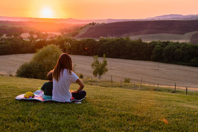 Rear view of mother sitting with son on field during sunset