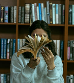 Young woman holding book against shelf in library