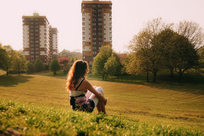 Woman sitting on grassy field by buildings against clear sky