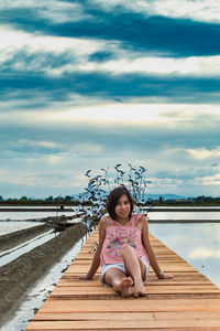 Portrait of mid adult woman sitting on pier over lake against cloudy sky during sunset