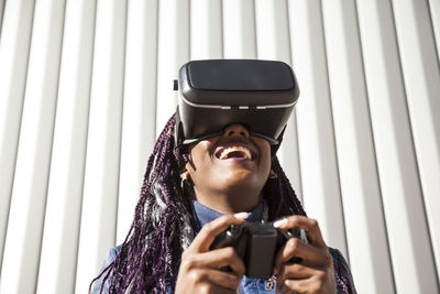 Excited young african american female in vr headset using controller while entertaining and playing virtual game against gray striped wall
