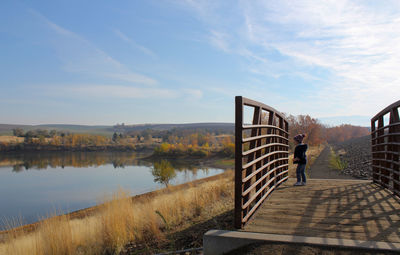 Side view of child standing on footbridge by lake against sky