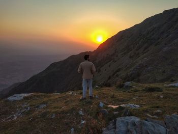 Rear view of man standing on rock by mountain against sky during sunset