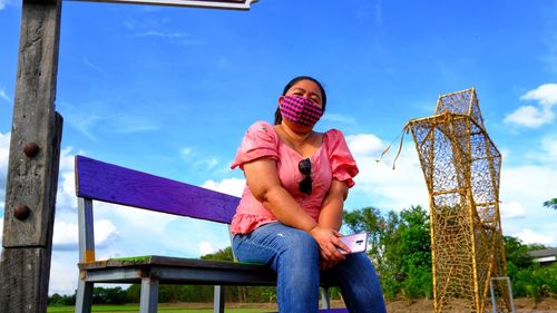 Portrait of woman wearing mask while sitting on bench against sky