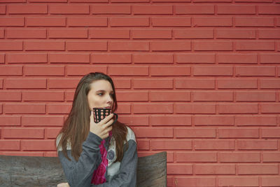 Thoughtful young woman holding cup against brick wall