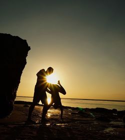 Silhouette man helping friend handstand at beach against sky during sunset