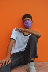 A indian young guy wearing face mask, looking at camera sitting against orange wall with copy space 