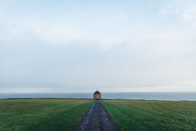 Footpath on grassy field leading towards building by sea against sky