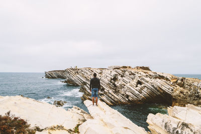 Rear view of man standing on cliff by sea against sky