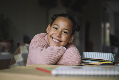 Portrait of happy girl resting head on arms while sitting on table with colored pencils at home