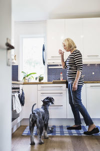 Senior woman drinking water while standing by dog in kitchen