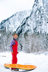 Portrait of girl in winter clothes with sleight standing against snow covered mountain