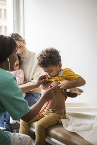 Female doctor examining boy's heartbeat with stethoscope while mother sitting and daughter in clinic