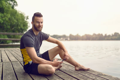 Full length portrait of smiling handsome man sitting on boardwalk by lake