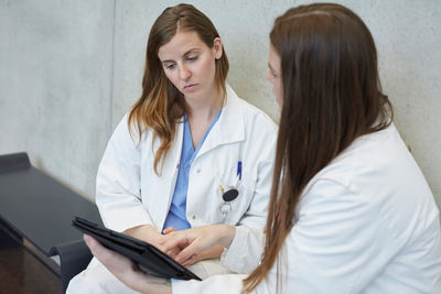 Confident young female doctor discussing with coworker over digital tablet while sitting against wall at hospital