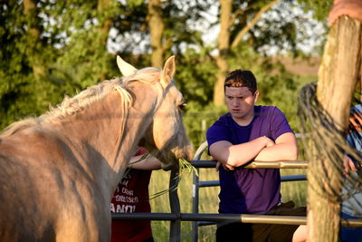 Young man looking at horse while standing by railing against trees