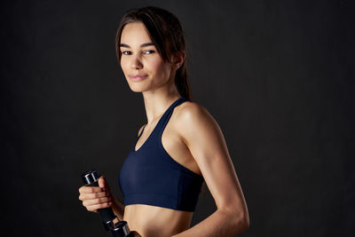 Portrait of young woman holding dumbbells while standing against black background