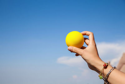 Cropped hand holding yellow ball against sky