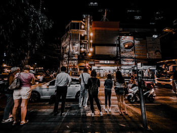 Rear view of people standing on illuminated street against buildings at night