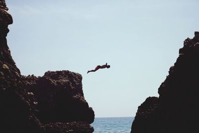 Man jumping on rock by sea against sky