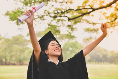 Young woman in graduation gown with arms raised holding certificate while standing at park