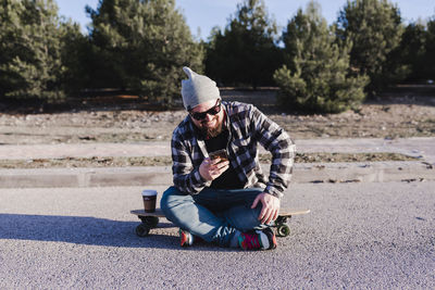 Modern young man sitting on a longboard looking at his mobile phone