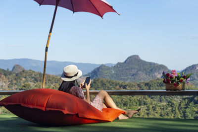 Rear view of woman wearing hat and umbrella sitting against landscape while using smart phone