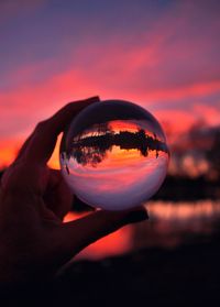 Reflection of person hand holding crystal ball against sky during sunset