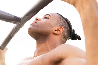 Low angle side view of focused shirtless hispanic male athlete doing pull ups on bar during fitness workout in park in daytime