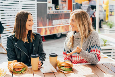 Two happy hungry female friends are chatting and eating burgers at the street market.