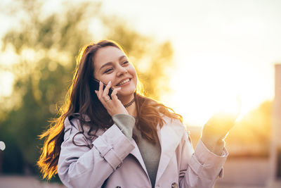 Smiling beautiful girl with long hair in a grey trench coat using smartphone in the spring 