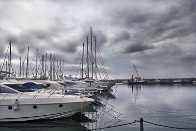 Sailboats moored at harbor against cloudy sky