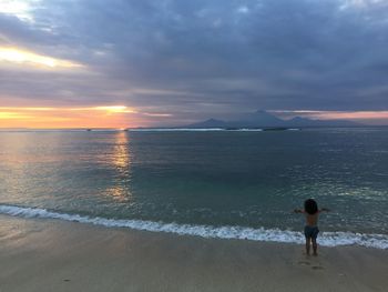 Rear view of shirtless child standing on shore at beach against cloudy sky during sunset