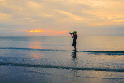 Side view of man photographing from dslr camera on shore against sky during sunset
