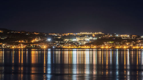 Illuminated buildings by river against sky at night