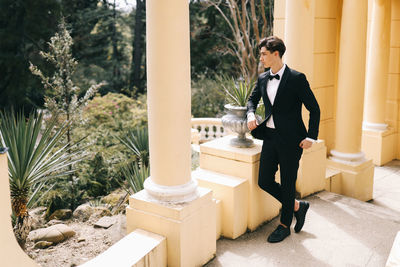 A beautiful young man, the groom in an elegant wedding suit, stands posing in the city's old park