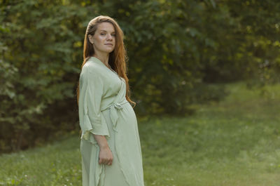 White caucasian pregnant woman stands, looks at camera. green trees, park is on background. 