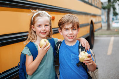 Children boy and girl students friends eating apples healthy snack by yellow school bus outdoors.