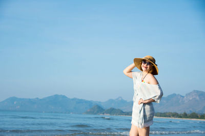 Smiling woman wearing sunglasses and hat against blue sky at beach