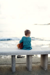 Cute european baby toddler in a green dress with a doll sits on a stone bench by  sea