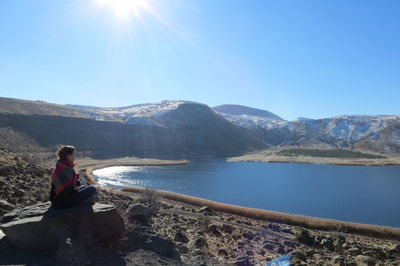 Man sitting on mountain by sea against sky on sunny day