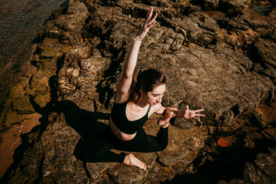 From above of flexible female sitting on stony shore near river in eka pada rajakapotasana and practicing mindfulness while doing yoga and looking away